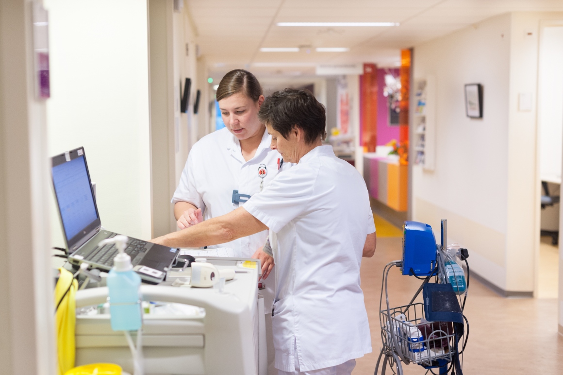 Twee verpleegkundigen overleggen op een gang bij een laptop op wielen over duurzaamheid.